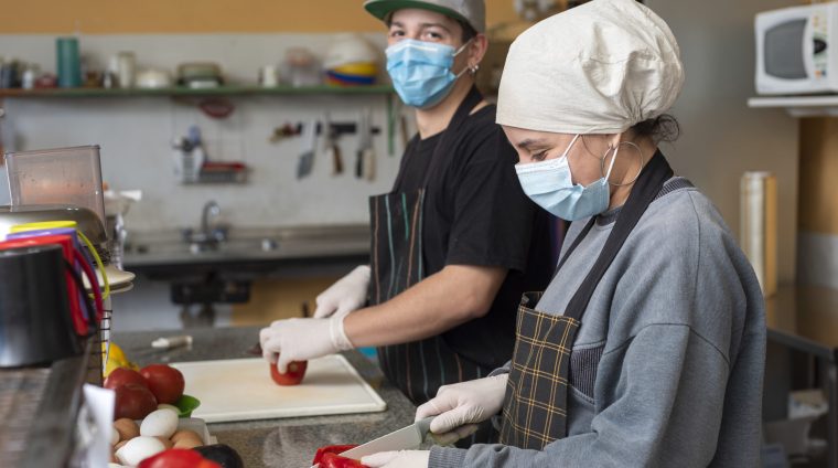 The two young chefs slicing vegetables wearing medical masks